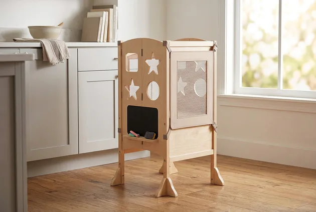 Natural wood Guidecraft Classic Kitchen Helper tower with star cutouts beside kitchen cabinets